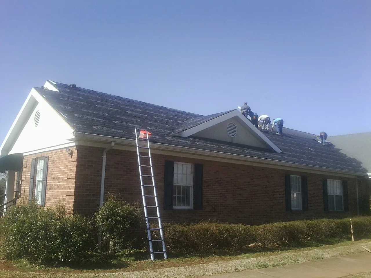 Crew tearing shingles off a brick ranch roof with a ladder set — Emergency Roof Repair work by our crew in Okemos